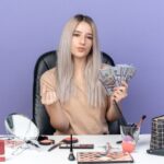 confident-young-beautiful-girl-sits-table-with-makeup-tools-holding-cash-showing-tip-gesture-isolated-blue-wall_141793-105686